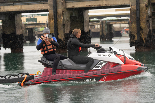 Two people ride a red Yamaha jet ski; one drives while the other uses the Outex Camera Pro Kit to take photos near a dock.