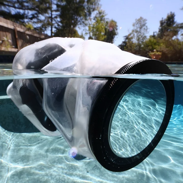 A camera in a clear waterproof housing is half-submerged in a pool, with sunlight reflecting off the water and trees visible in the background.