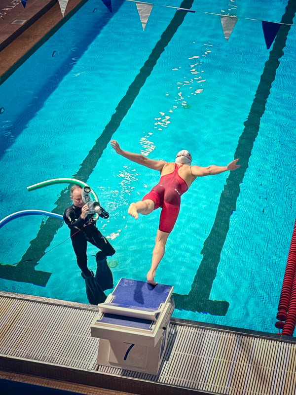 A behind-the-scenes overhead view of photographer Finn O’Hara in a wetsuit, submerged in a swimming pool while using an Outex underwater housing to photograph an athlete diving off the starting blocks.