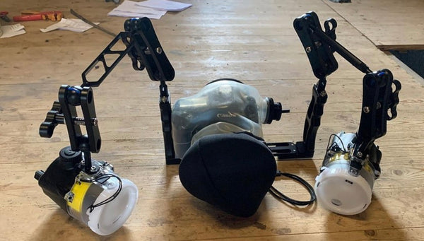 An Outex Flash, Data, or Tripod Add-on Kit with underwater strobes and adjustable arms sits on a wooden floor, featuring a lens cover in front.