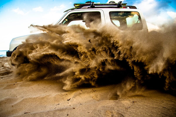 A white SUV drives through sand on a beach, kicking up a large cloud of sand and dust in front of its wheels.