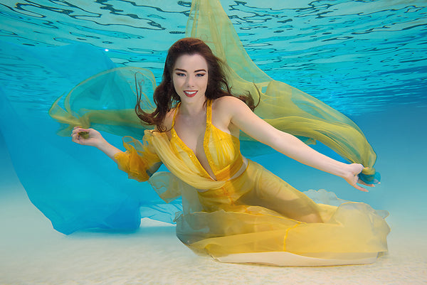 A woman with her arms spread in a yellow dress is inside a pool with blue water