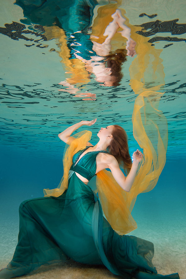 A woman with her arms spread in a green dress is underwater in a pool with blue water