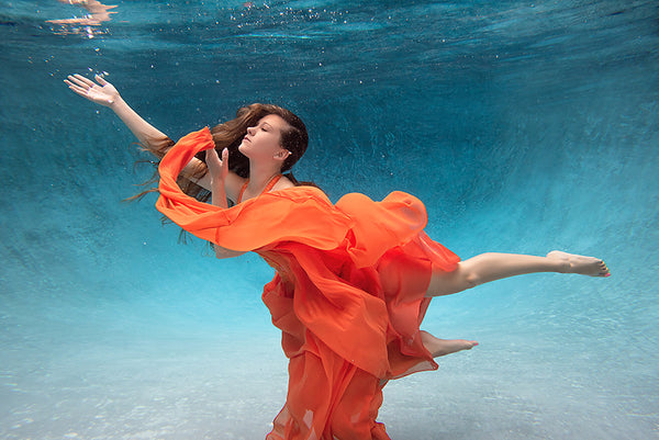 A woman with her arms spread in a red dress is underwater in a pool with blue water