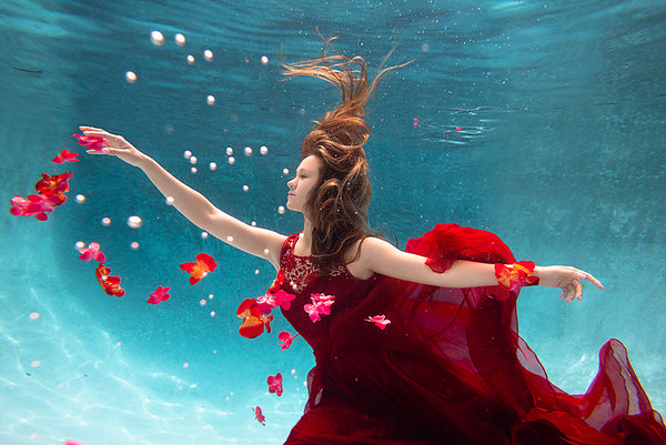 A woman with her arms spread in a red dress is underwater in a pool with blue water