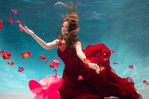 A woman with her arms spread in a red dress is underwater in a pool with blue water