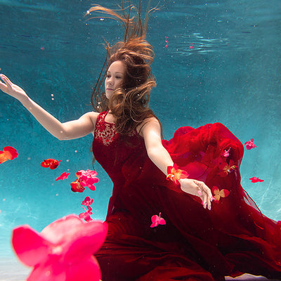 A woman with her arms spread in a red dress is underwater in a pool with blue water