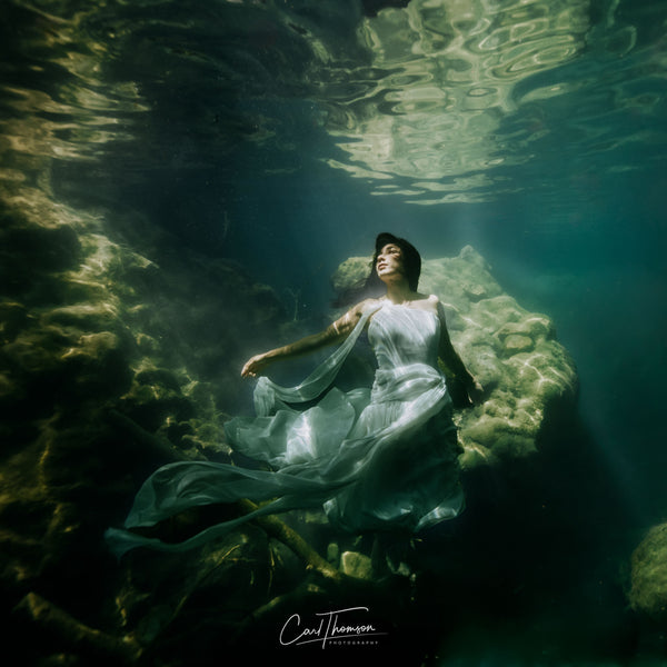 Underwater portrait of a woman in a flowing white couture dress floating among submerged rocks in a French river, photographed by Carl Thomson using an Outex underwater housing on a Nikon D850