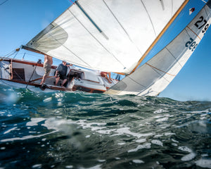Sailboat with people onboard tilting on choppy blue water under clear sky, with white sails catching the wind.