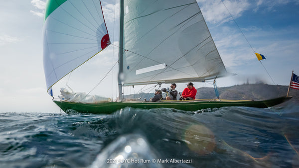 A sailboat with three people glides over ocean waves under a partly cloudy sky, with mountains and flags visible in the background.