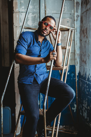 Man in red glasses and blue shirt sits on metal stairs in an industrial setting, leaning on the railing and looking at the camera.