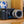 A black GoPro camera with a silver lens, resting on a wooden shelf in front of a wooden panel, with decorative ceramic mugs in the background.