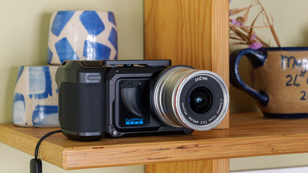 A black GoPro camera with a silver lens, resting on a wooden shelf in front of a wooden panel, with decorative ceramic mugs in the background.