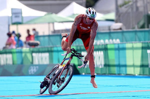 A triathlete in a red suit dismounts his bike mid-air during a race, about to land on a blue track, with event banners in the background.