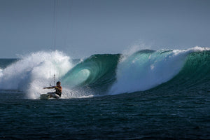 A person kite surfing on the ocean, riding a wave with water splashing around under a clear blue sky.