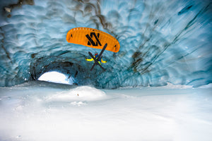 A skier with a yellow parachute glides through a blue ice cave, soaring above snow-covered ground inside the tunnel.