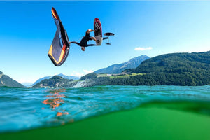 A person performs an aerial trick on a wing foil above green water, with mountains and blue sky in the background.