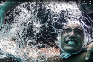 A person underwater grins widely, surrounded by bubbles and swirling water, wearing a swim cap with chin straps.