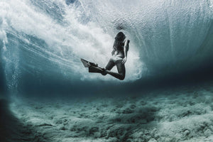 A person with fins swims underwater beneath a breaking wave, surrounded by swirling bubbles and a sandy ocean floor.