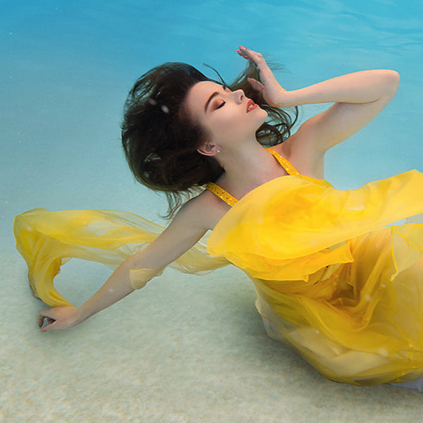 A woman with her arms spread in a yellow dress is inside a pool with blue water