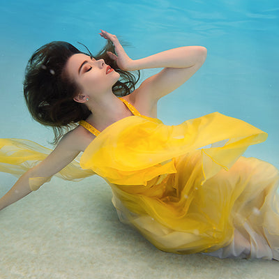A woman with her arms spread in a yellow dress is inside a pool with blue water