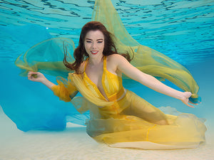 A woman with her arms spread in a yellow dress is inside a pool with blue water