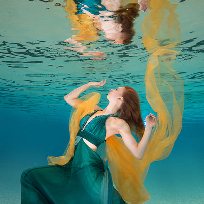 A woman with her arms spread in a green dress is underwater in a pool with blue water