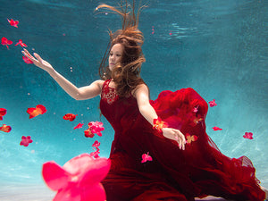 A woman with her arms spread in a red dress is underwater in a pool with blue water